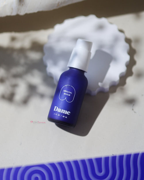 A bottle of Dame's Arousal Serum with a blue label, placed on a surface with a white patterned background.